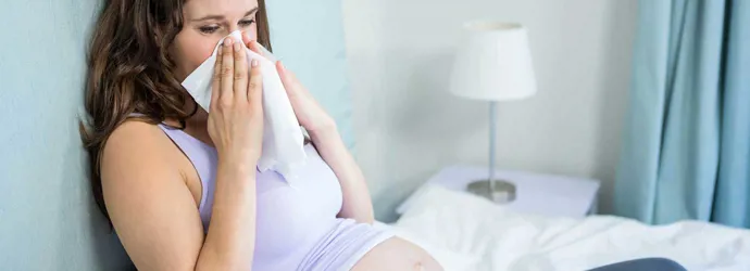 A pregnant woman sat on a bed blowing her nose into a tissue