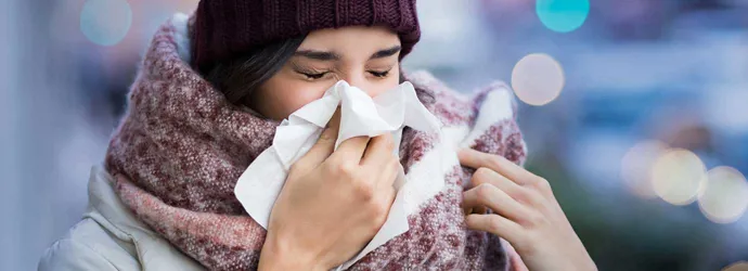Woman wrapped up in bulky winter wear suffering from sinusitis symptoms and sneezing into a tissue outdoors