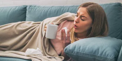Woman lying in the sofa coughing while holding tea cup and a tissue
