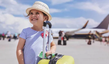 Young girl smiling with a plane in the background holding a yellow suitcase of items you need for a family holiday
